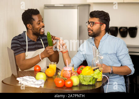 Two students argue what to cook for dinner. Multicultural friends sitting in kitchen, using laptop computer talking and preparing vegetable salad, smo Stock Photo