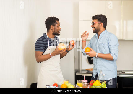 Cheerful indian young man in glasses and african american man wearing apron having fun in kitchen. Two male friends standing near desk with fruits and Stock Photo