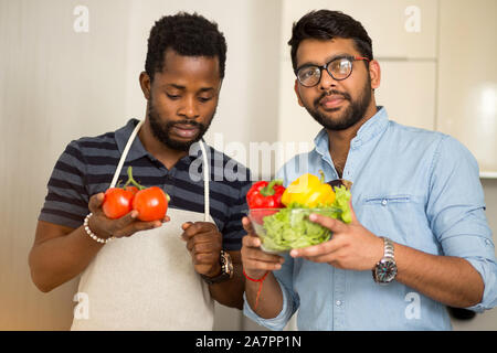 Closeup shot of two male chef friends standing in light kitchen and holding fresh organic vegetables and looking at camera. Healthy food, farm product Stock Photo