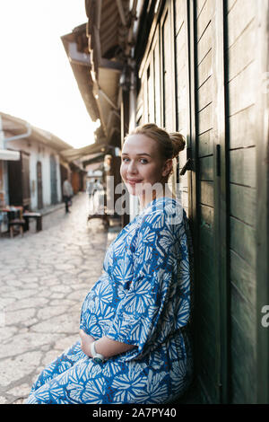 Portrait of beautiful pregnant woman. She is standing in the middle of the street and holding hands on her belly. Stock Photo