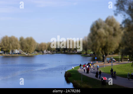 MUNICH, BAVARIA, GERMANY – OCTOBER 27, 2019: Tilt-shift of people walking on a walking path at the shore of the Olympiasee pond in Olympiapark Stock Photo