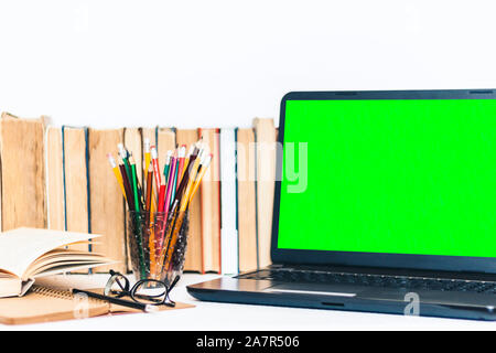 Green screen laptop, pile if books, notebook, smartphone, glasses and pencils in holder on white table, education concept background Stock Photo