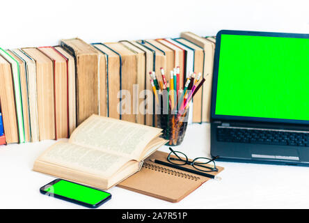 Green screen laptop and smartphone, pile if books, notebook,  glasses and pencils in holder on white table, education concept background Stock Photo