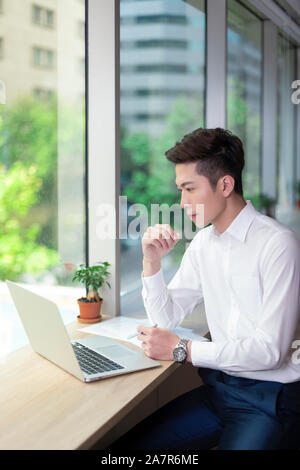 Waist up shot of a busy smiling young businessman with short black hair and a white button down shirt using a laptop at a desk in an office Stock Photo