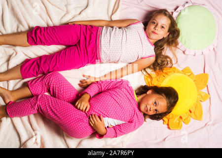 Children with smiling faces lie close on light pink blanket background. Childhood, party and happiness concept. Kids in pink pajamas have fun. Schoolgirls having pajama party with funny pillows. Stock Photo