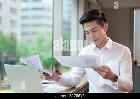 Waist up shot of a busy smiling young businessman with short black hair and a white button down reading documents at a desk in an office Stock Photo