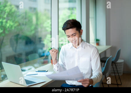 Young businessman with short black hair and a white button down shirt showing a success gesture while reading a document at a desk in an office Stock Photo