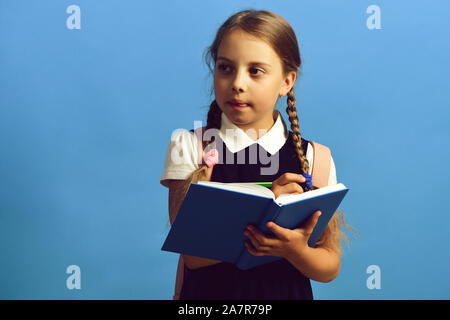 Girl writes in big blue notebook. Pupil in school uniform with braids. School girl with thoughtful face expression having idea, blue background. Back to school and education concept Stock Photo