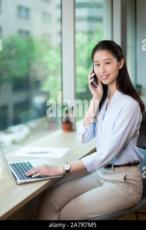 Smiling young businesswoman with long straight black hair using a smartphone and a laptop while looking at the camera at a desk in an office Stock Photo