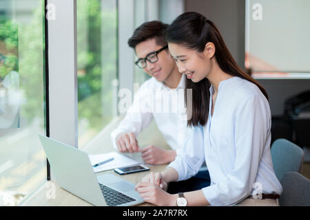 Waist up shot of two smiling young male and female businesspeople with black hair using a laptop together while working at a desk in an office Stock Photo