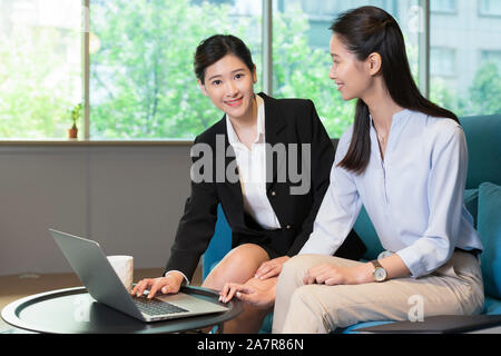 Photograph of two young smiling businesswomen using a laptop on a sofa in an office with one of them looking at the camera Stock Photo