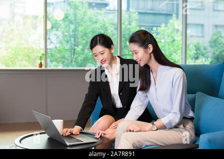 Photograph of two young smiling businesswomen using a laptop together on a blue sofa in an office Stock Photo
