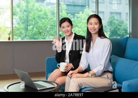 Photograph of two young businesswomen sitting on a blue sofa in an office and smiling at the camera with one also waving Stock Photo