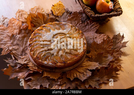 Freshly baked homemade apple pie with cup of tea near basket with apples on wooden table and autumn yellow and red foliage. Stock Photo