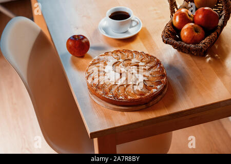 Freshly baked homemade apple pie with cup of tea near basket with apples on wooden table. Minimalism Stock Photo