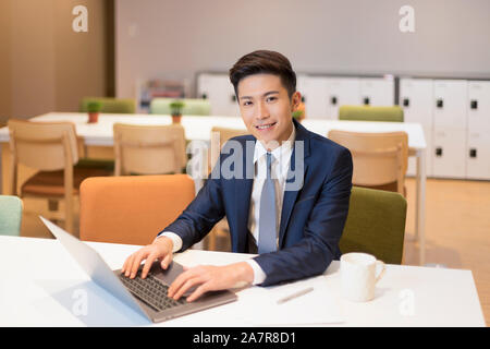 Waist up portrait of a young smiling businessman in a suit sitting at a desk in an office in front of a laptop and looking at the camera Stock Photo