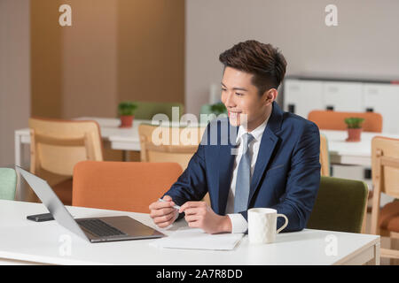 Waist up shot of a smiling young smiling businessman in a suit sitting at a desk in an office in front of a laptop and working Stock Photo