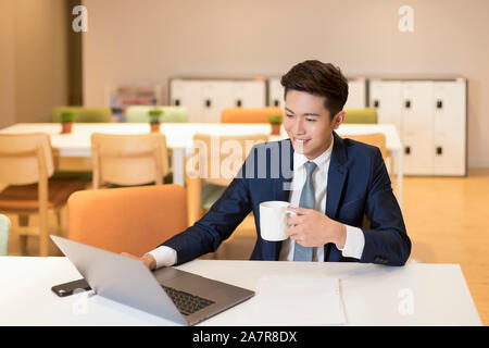 Waist up shot of a smiling young smiling businessman in a suit sitting at a desk in an office in front of a laptop and drinking coffee and working Stock Photo
