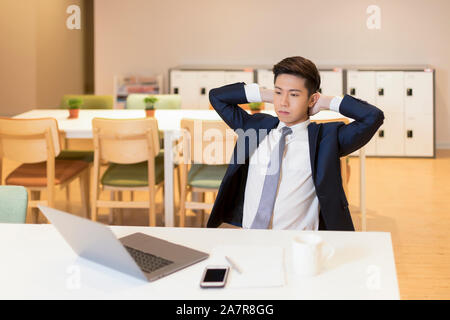 Waist up shot of a young businessman with black hair and suit sitting at a desk in an office with a serious expression and hands behind head Stock Photo