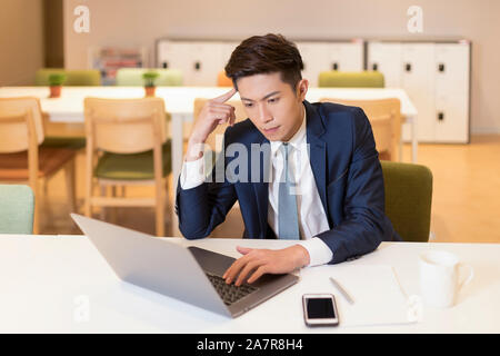 Waist up shot of a young businessman with black hair and a suit sitting concentrated on work in front of laptop at the desk in an office Stock Photo