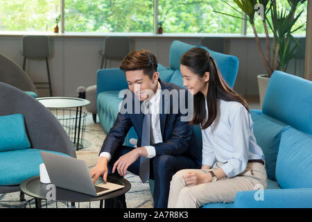 Photograph of two smiling young male and female businesspeople working at a laptop together while sitting on a blue sofa in an office Stock Photo
