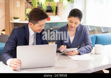 Photograph of two male and female young smiling businesspeople using a tablet and a laptop together while working in an office Stock Photo