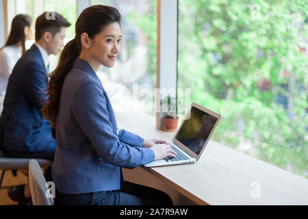 Side view shot of a smiling young businesswoman with a black ponytail using a laptop at a desk in an office Stock Photo