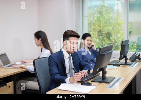 three young male and female businesspeople working in an office with one using computer, another one using phone and another one using laptop Stock Photo