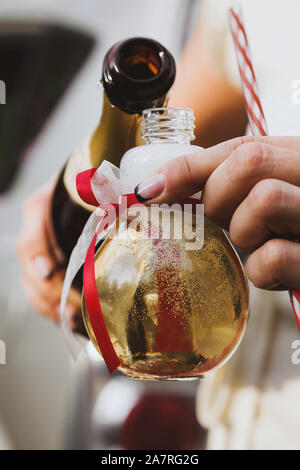 Female hands pouring champagne into a light bilb shaped party glass decorated with red and white ribbons Stock Photo