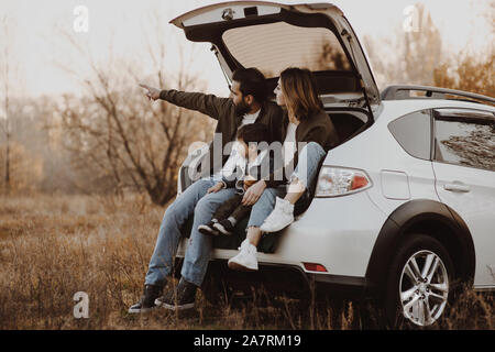 Happy family sitting at open trunk of hatchback car and pointing finger outdoors. Road trip concept. Stock Photo