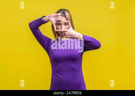 Portrait of concentrated woman in elegant tight purple dress looking at camera through hand frame, capturing photo or focusing eyesight on target. ind Stock Photo