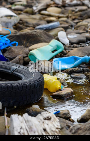 Enviromental pollution. Rubber tire with plastic bottles and waste in dirty water on ocean beach. Stock Photo