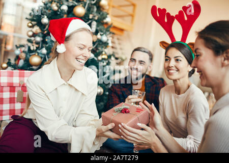 Group of cheerful adult people exchanging gifts during Christmas party, focus on happy young woman in foreground Stock Photo