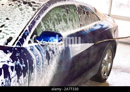 A car wash worker washes blue car with high-pressure apparatus. Foam. Rear side view. Stock Photo