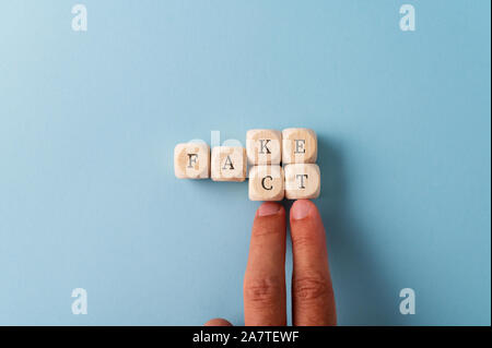 Word Fake spelled on wooden dices with male hand changing the last two letters to spell the word Fact. Over blue background with copy space. Stock Photo