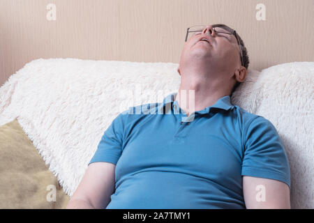 White middle-aged man fell asleep sitting on a sofa. A man with glasses and a blue T-shirt sleeps while sitting Stock Photo