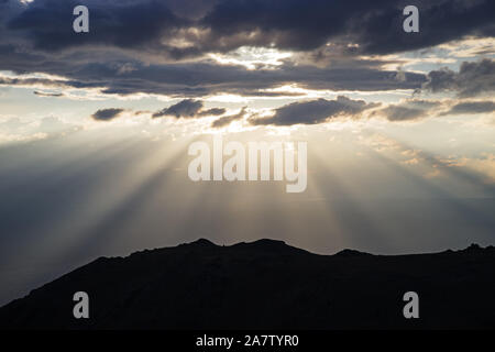 sunrays stream out from clouds behind a hill Stock Photo