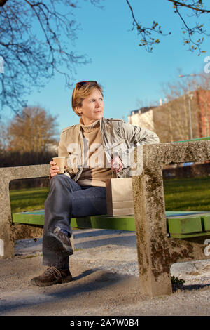 A woman seats on the bench in a park enjoying in the lunch break. Paper, eco cup and bag. Stock Photo