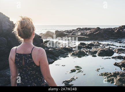 blonde woman in summer dress standing at rocky shore looking at ocean in sunlight Stock Photo