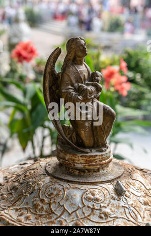 Da Nang, Vietnam - March 10, 2019: Closeup of bronze statue of winged angel with baby Jesus. Faded green and red of foliage and flowers. Stock Photo