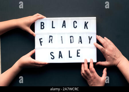 Lightbox text "Black Friday Sale" on black paper background. Creative flat lay with two pairs of hands, child and adult, holding and touching the ligh Stock Photo