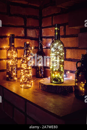 Lot of different vintage style beer bottles and green wine bottles decorated with wire string micro led lights on shelf, red brick wall background. Il Stock Photo