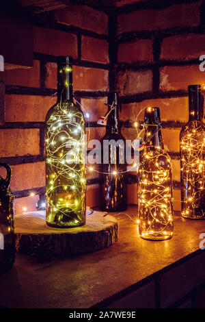 Lot of different vintage style beer bottles and green wine bottles decorated with wire string micro led lights on shelf, red brick wall background. Il Stock Photo