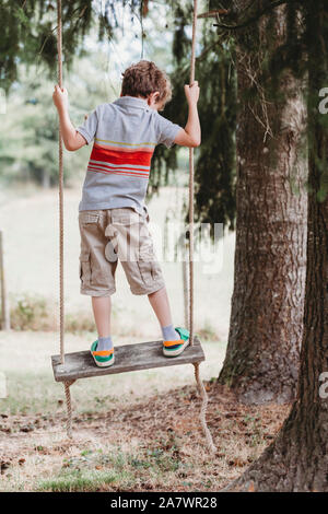 Rear view of boy standing on swing under pine trees Stock Photo