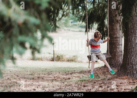 Front view of boy sitting on swing under pine trees Stock Photo