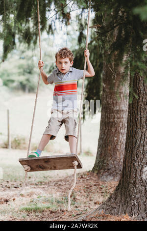 Vertical front view of boy standing on swing under pine trees Stock Photo