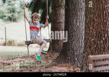Boy sitting sideways on swing under pine trees Stock Photo