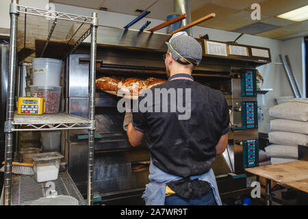 rear view of a baker removing loaves of bread from commercial oven Stock Photo