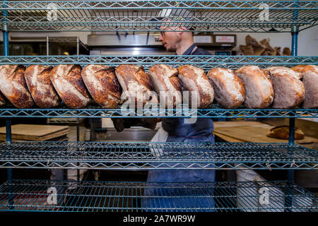 A baker walks by a rack filled with a line of fresh-baked bread loaves Stock Photo