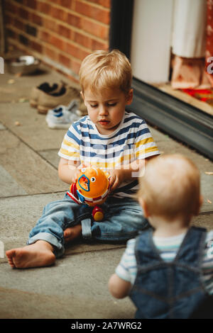 Toddler sitting outside playing with toy, watched by baby brother